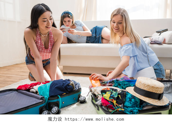 两个妇女在查看装满衣物的行李箱和一个妇女躺在沙发上看手机妇女包装箱子
